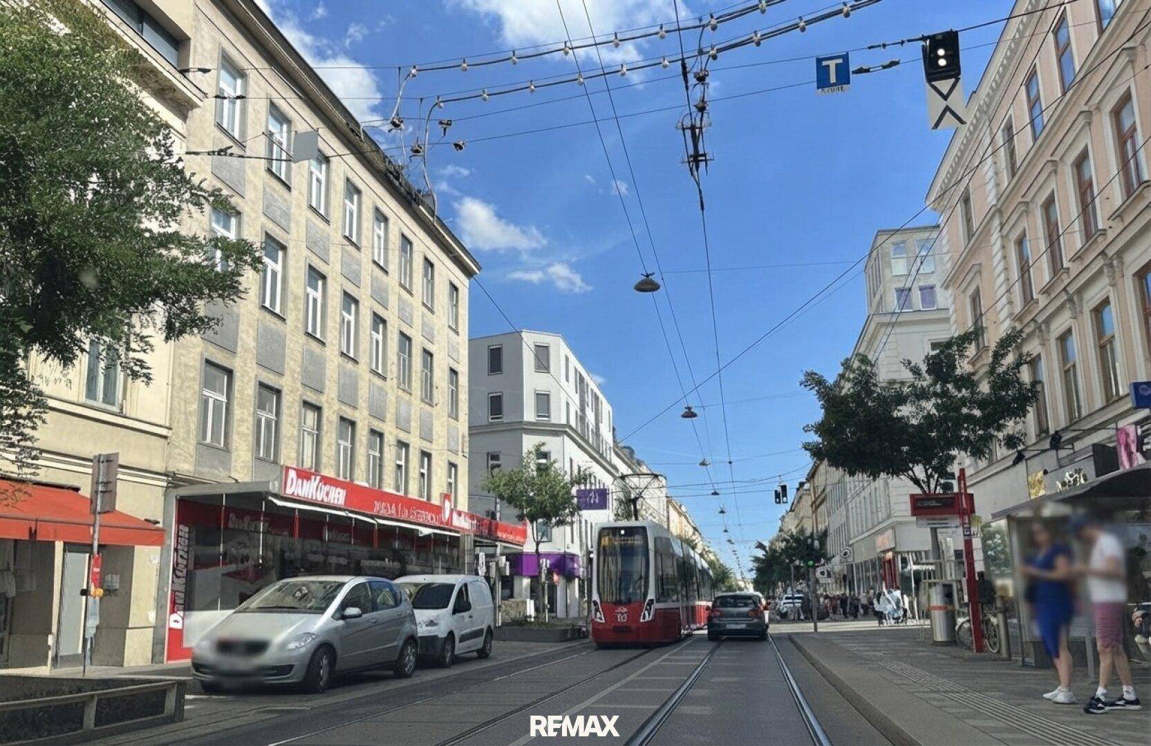 Belebtes Stadtbild mit Straßenbahn, Geschäften und Gebäuden unter blauem Himmel.