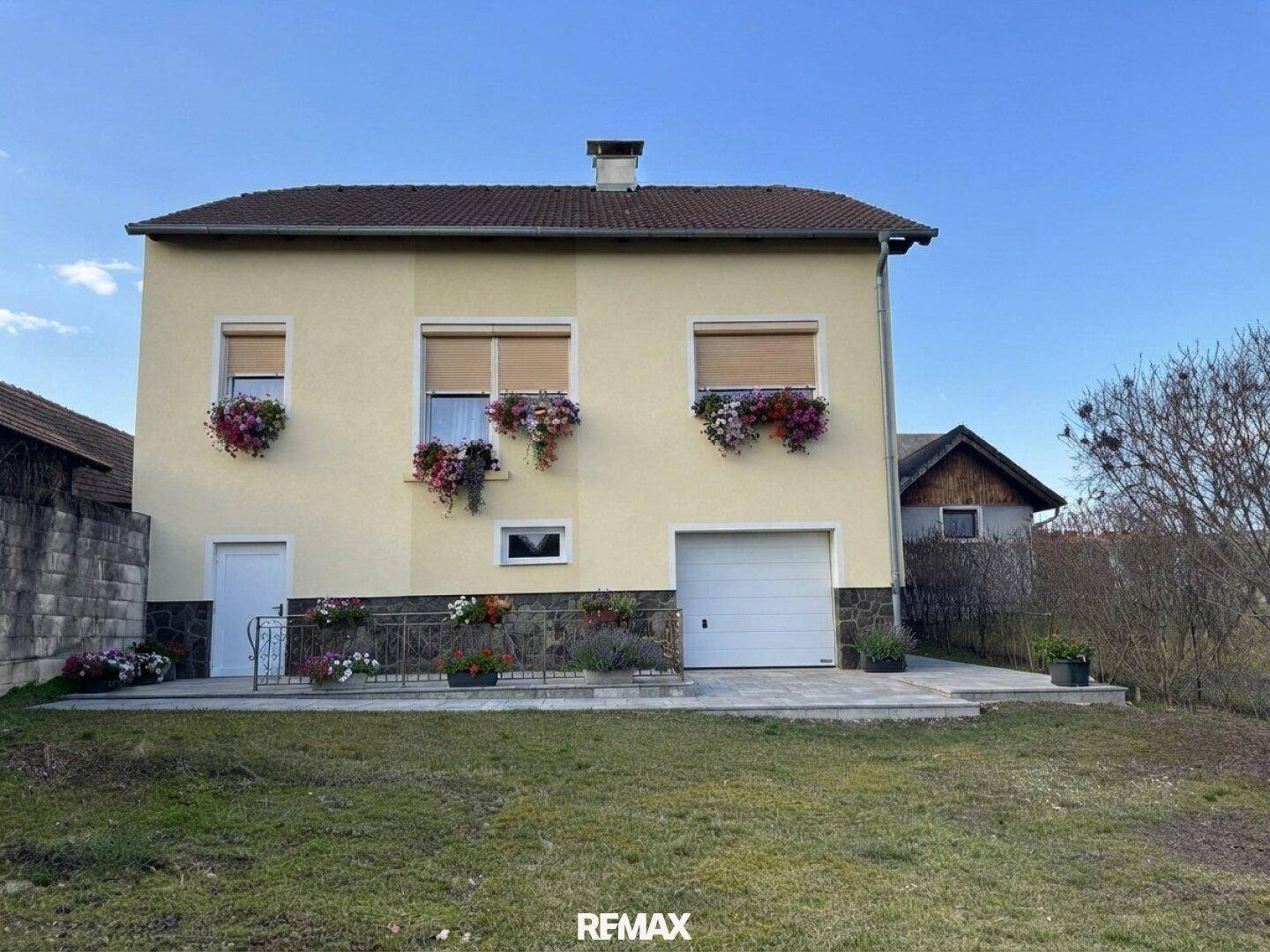 Gelbe Hausfassade mit Rollläden, Blumenkästen und integrierter Garage, umgeben von einem grünen Rasen.