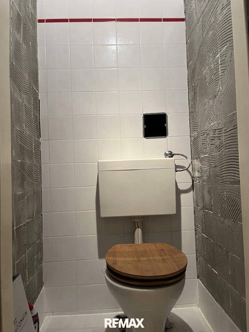 Kleines WC mit weißen Fliesen, roter Akzentlinie und einer älteren Toilette mit Holzdeckel.