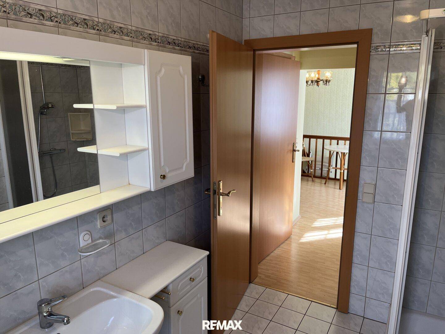 Badezimmer mit Waschbecken, Spiegelschrank und offener Tür zu einem weiteren Raum.