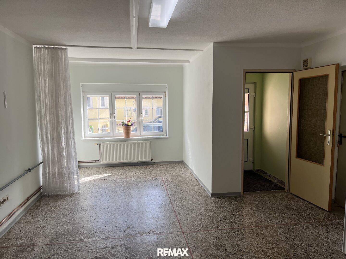 Geräumiger, heller Raum mit Terrazzoboden, großem Fenster und Zugang zum Flur.