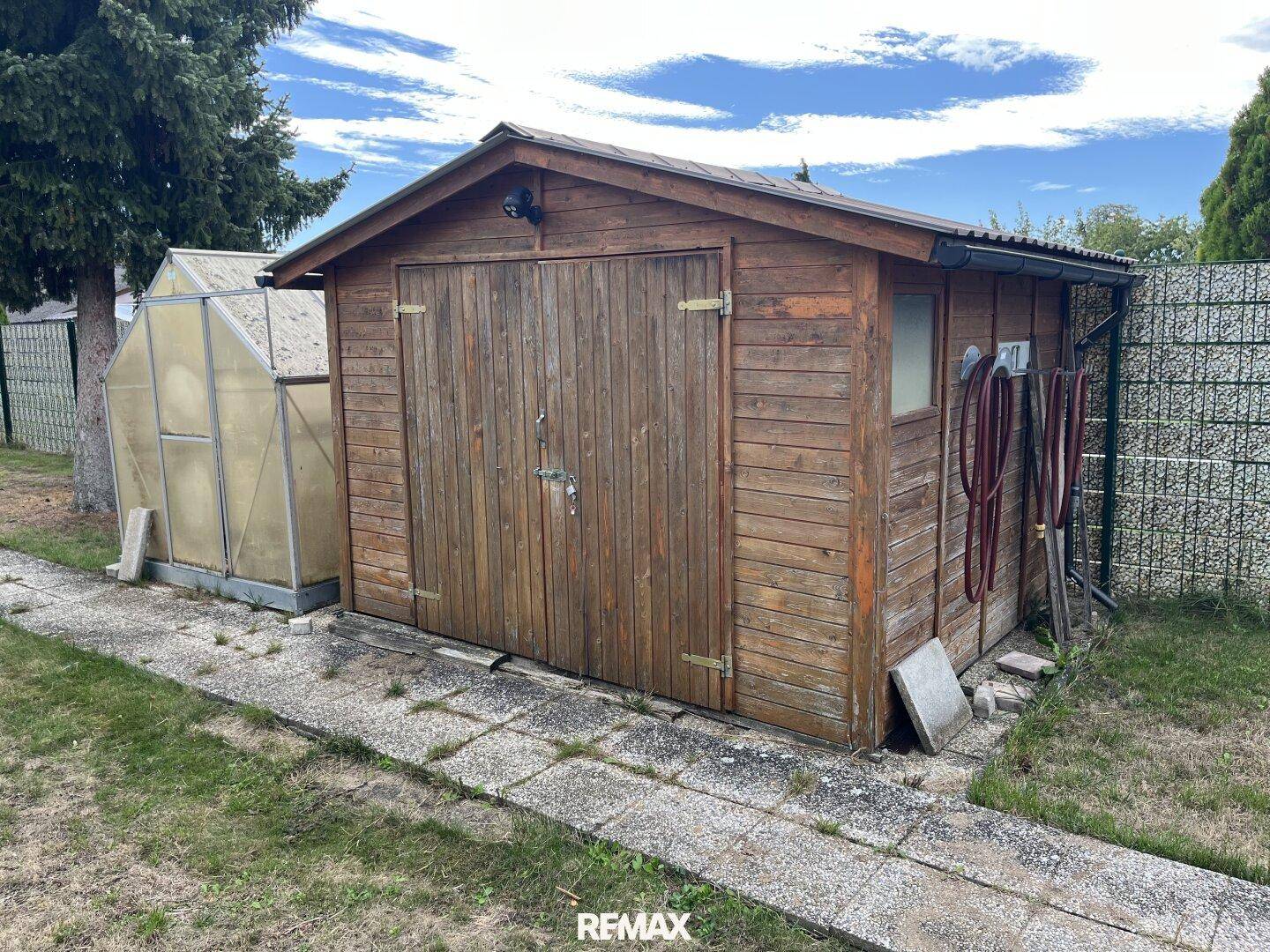 Rustikaler Holzschuppen im Garten mit angrenzendem Gewächshaus und gepflastertem Weg.