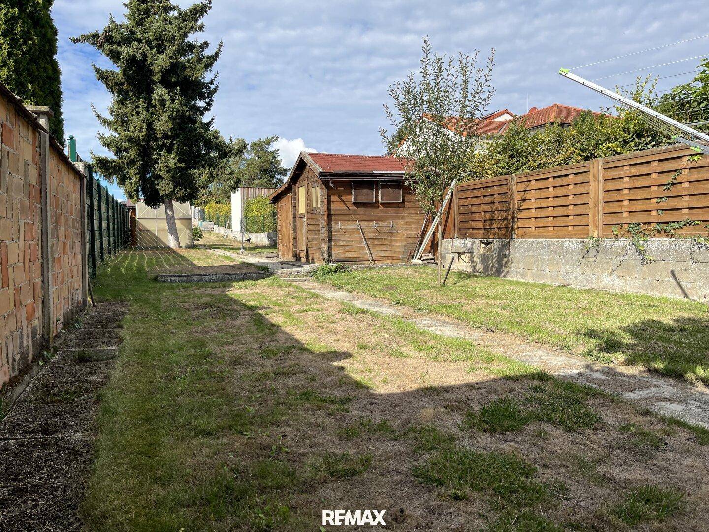 Langer Gartenbereich mit Rasenfläche, einem Holzschuppen im Hintergrund und seitlichen Zäunen.