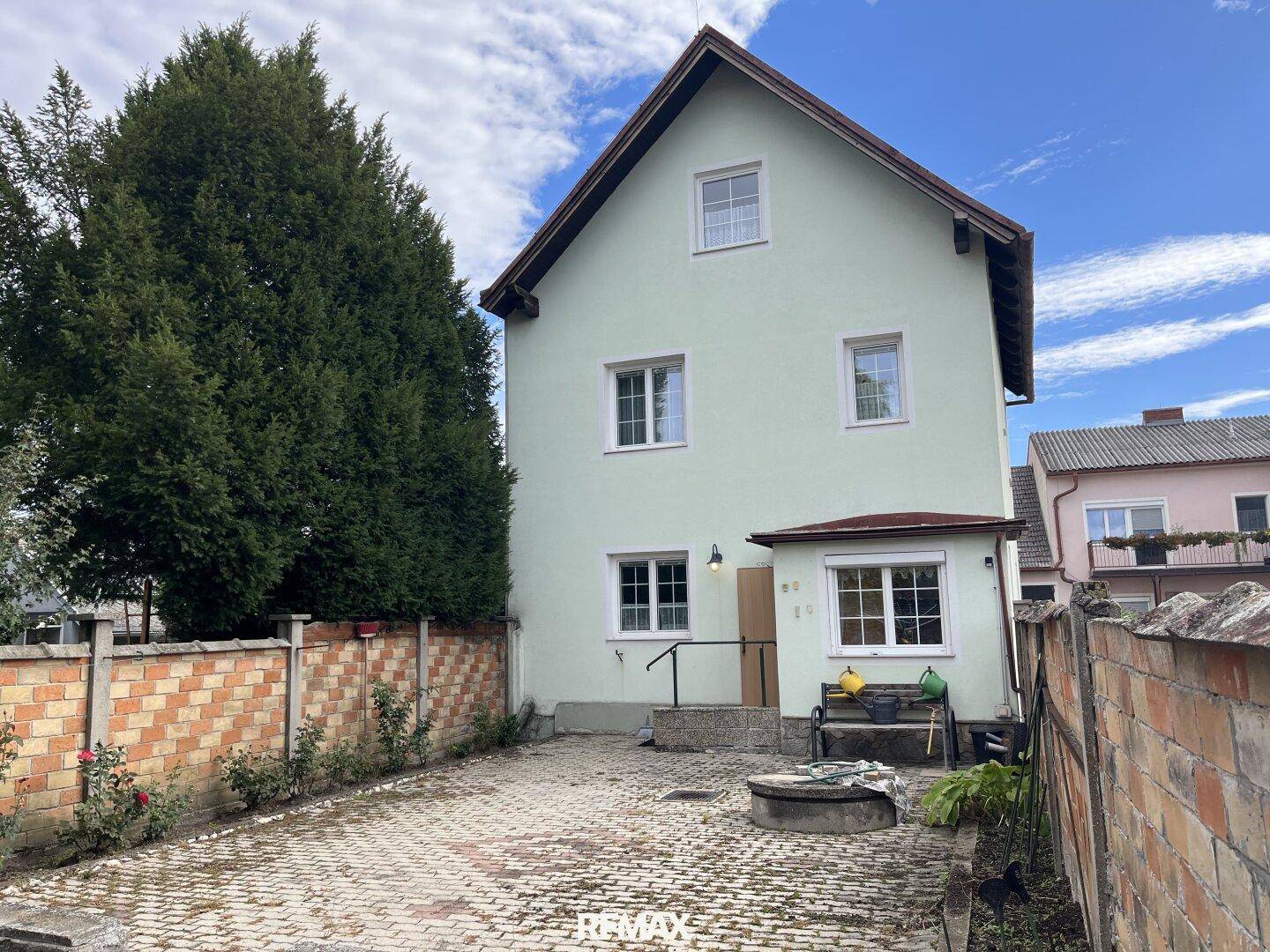 Hausfassade in hellem Grün mit weißen Fenstern und einem gepflasterten Vorplatz, umgeben von einer Ziegelmauer.