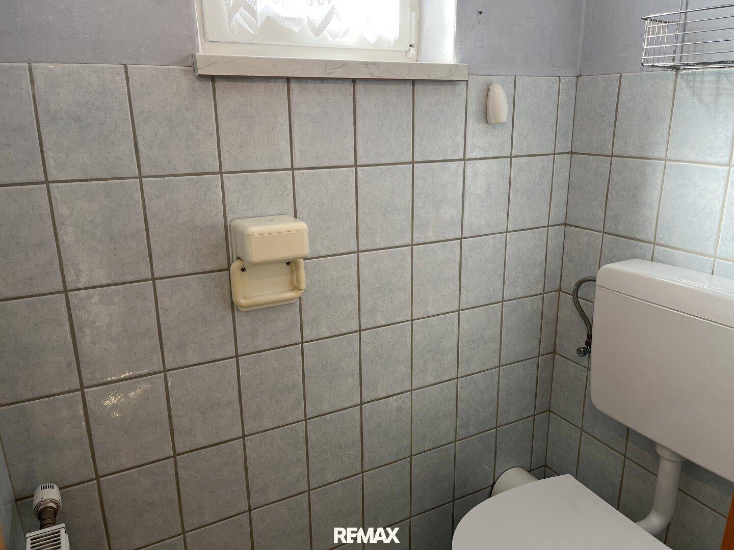 Kleines WC mit hellgrauen Fliesen, Toilette und Fenster für Belüftung.