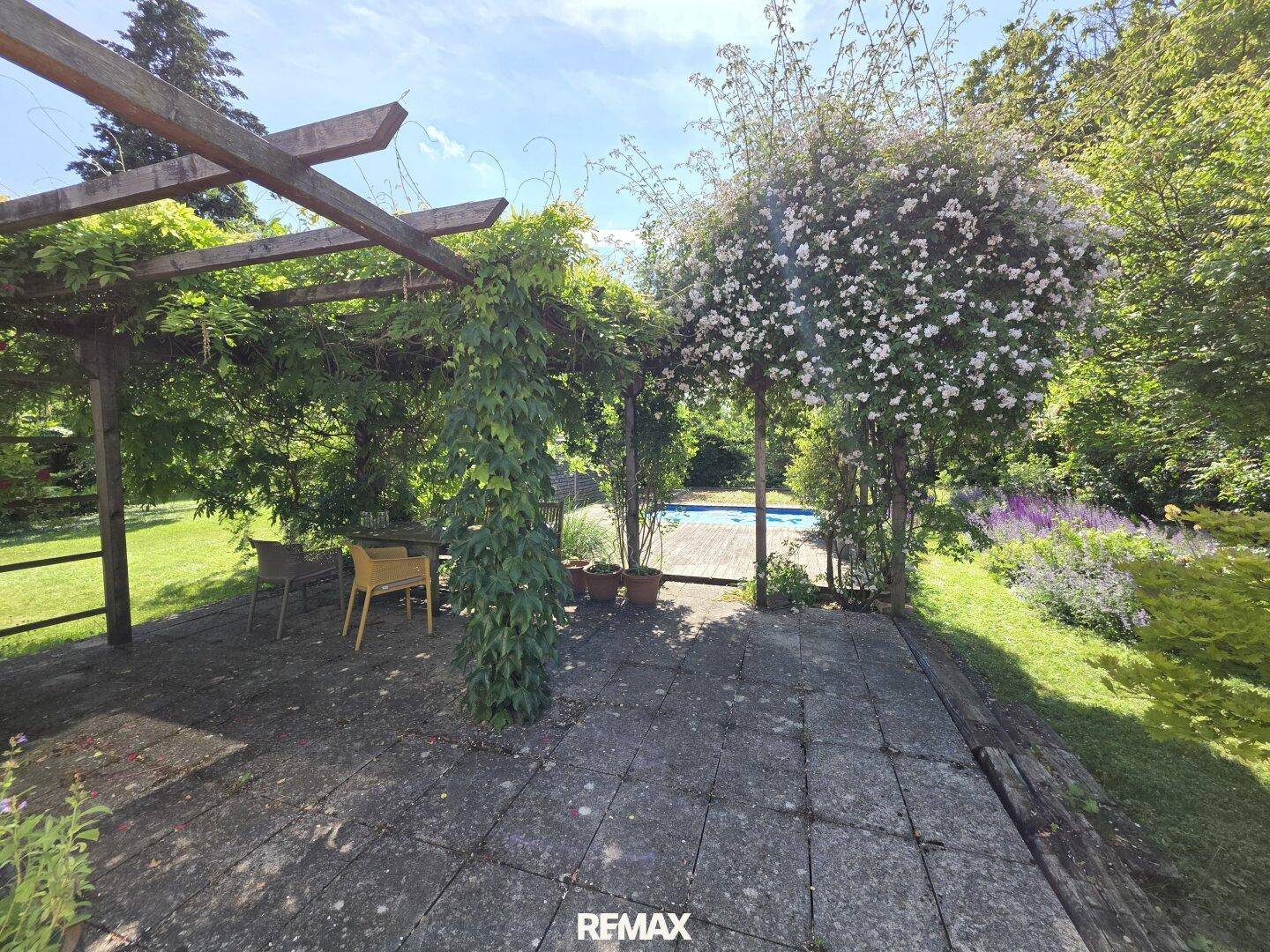Idyllischer Gartenbereich mit Pergola, Sitzgelegenheiten und einem Pool im Hintergrund, umgeben von üppigem Grün.