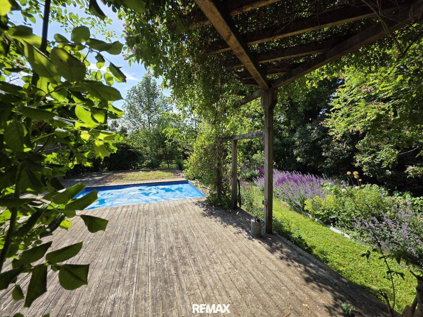 Große Holzterrasse im Garten mit Blick auf den Pool, umgeben von dichtem Laub und blühenden Pflanzen.