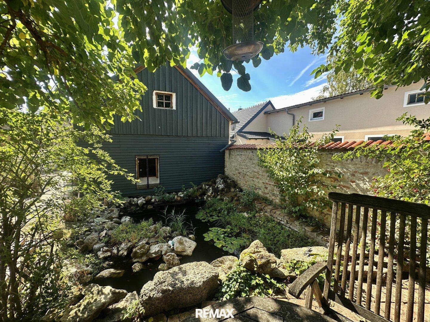 Idyllischer Gartenbereich mit Teich, umgeben von üppigem Grün und einem Holzhaus im Hintergrund.