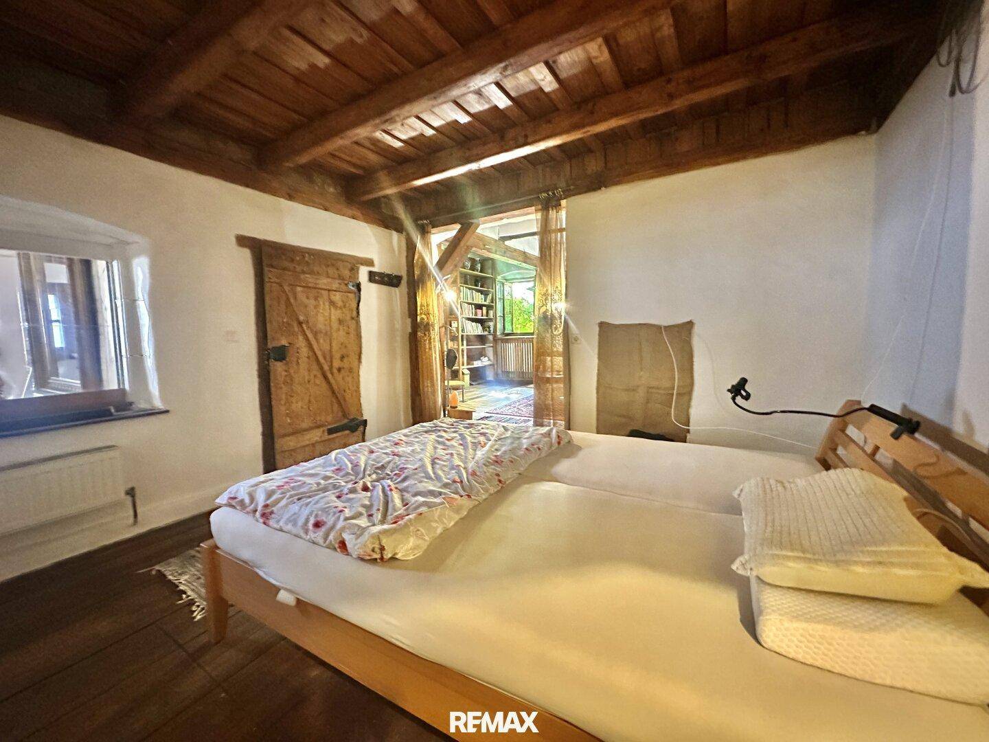 Schlafzimmer mit Holzbalkendecke, Holzboden und einem einfachen Bett, das zum Wohnbereich führt.