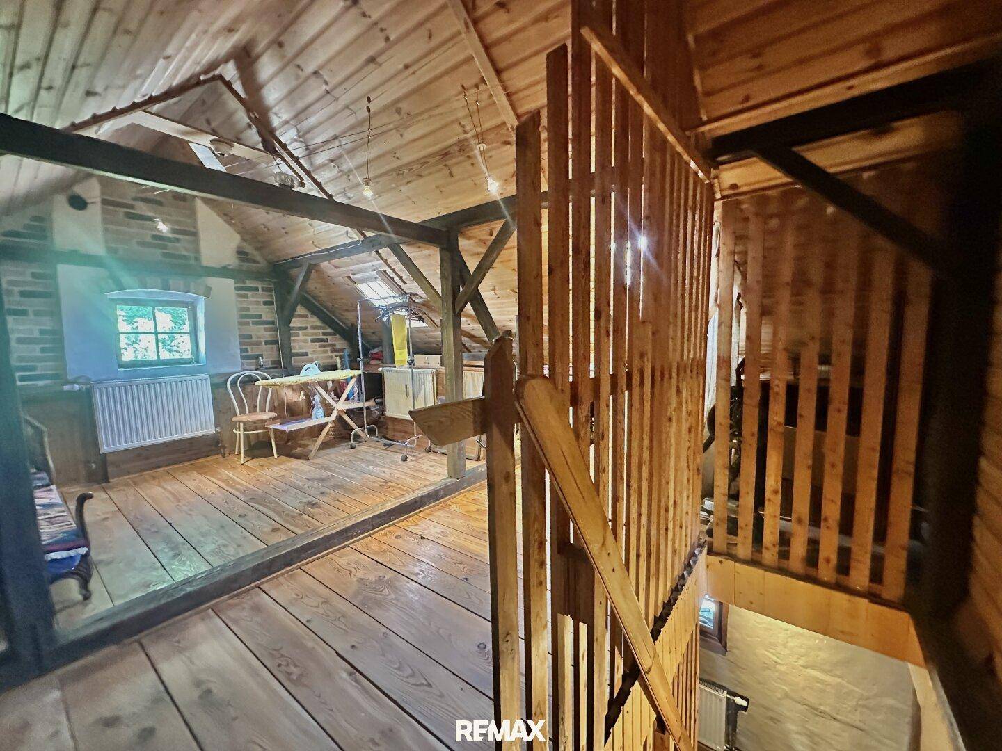 Ausgebauter Dachboden mit Holzbalken, Holzboden und einem kleinen Fenster, das Licht hereinlässt.