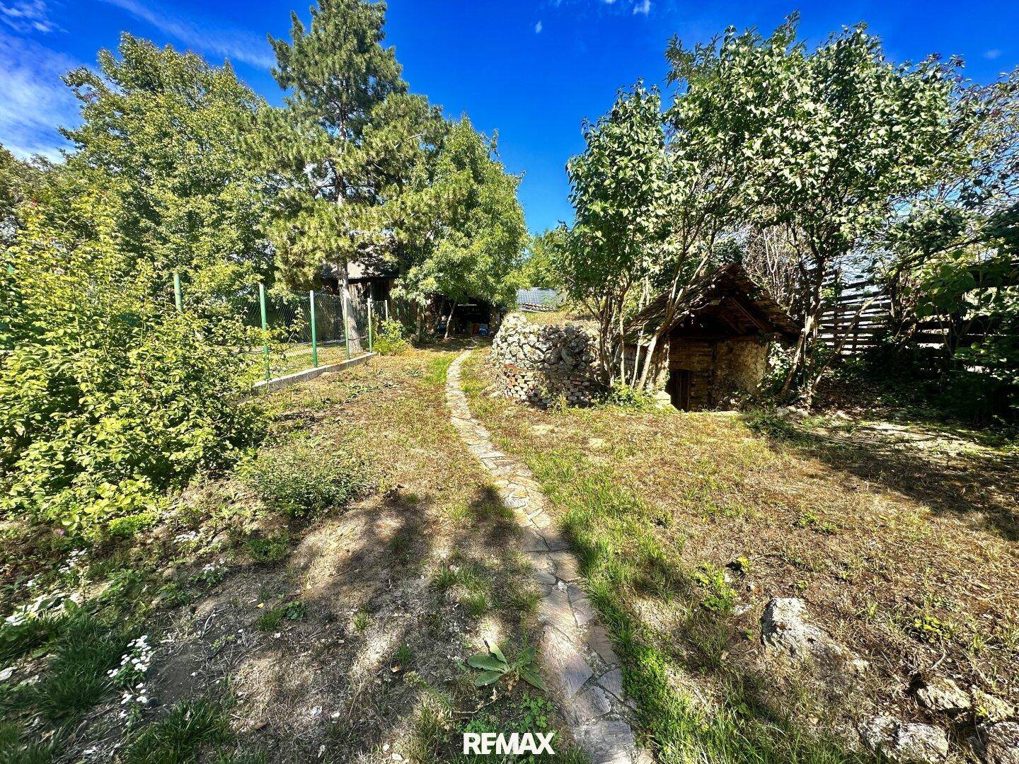 Weitläufiger Garten mit Steinpfad und alten Bäumen unter strahlend blauem Himmel.