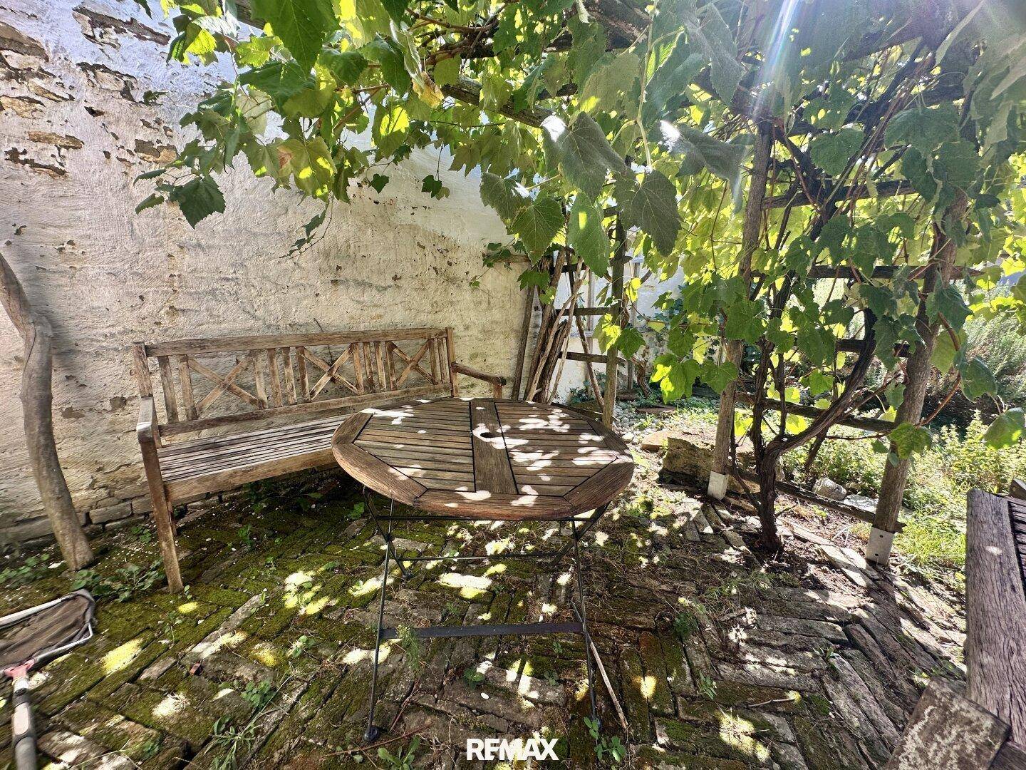Gemütlicher Außenbereich mit Holztisch und Bank unter schattenspendenden Bäumen.
