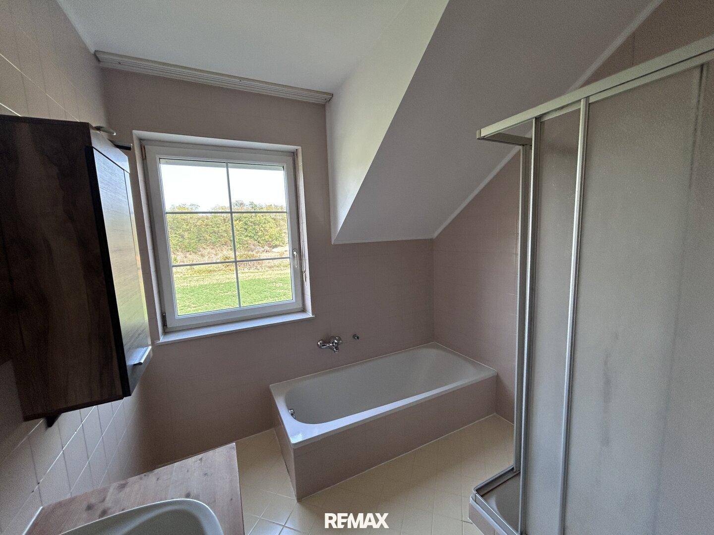 Badezimmer mit Badewanne, Duschkabine und Fenster, ausgestattet mit hellrosa Fliesen und Dachschräge.