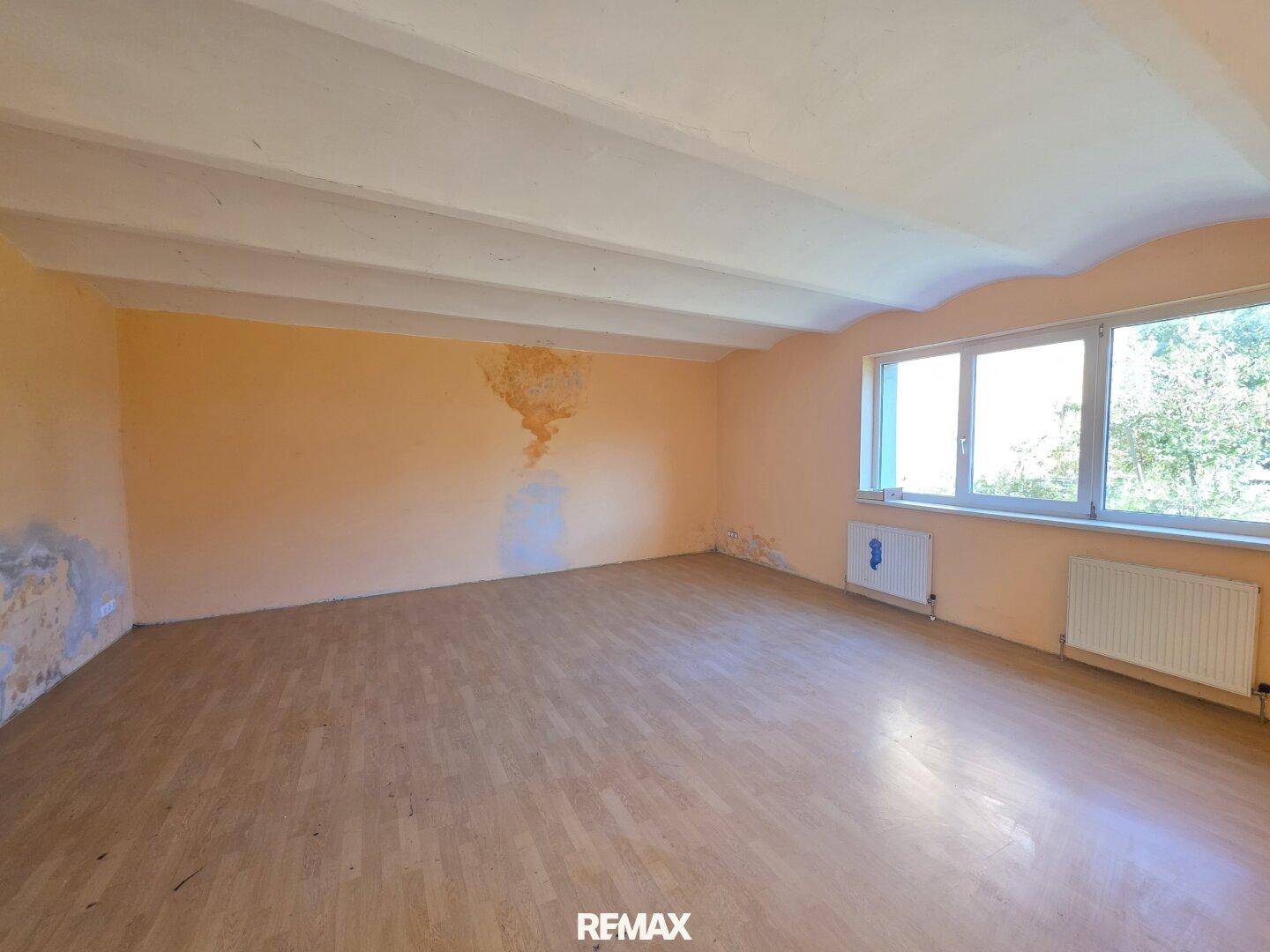 Geräumiger Raum mit Holzboden und großen Fenstern, der Renovierungsbedarf zeigt.