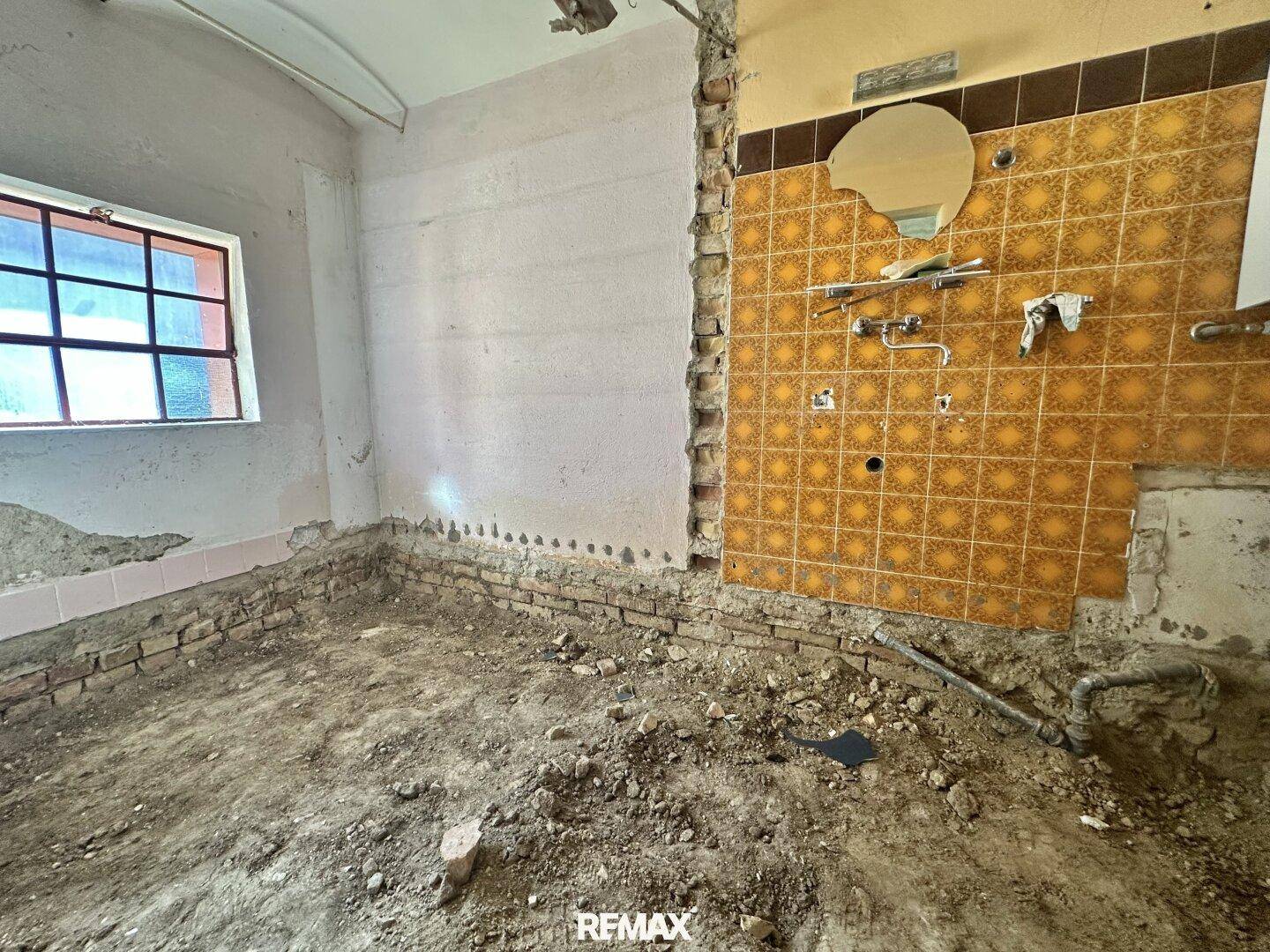Küche mit alten gelb-braunen Fliesen an der Wand, sichtbaren Rohren und unfertigem Boden, die eine Modernisierung benötigt.
