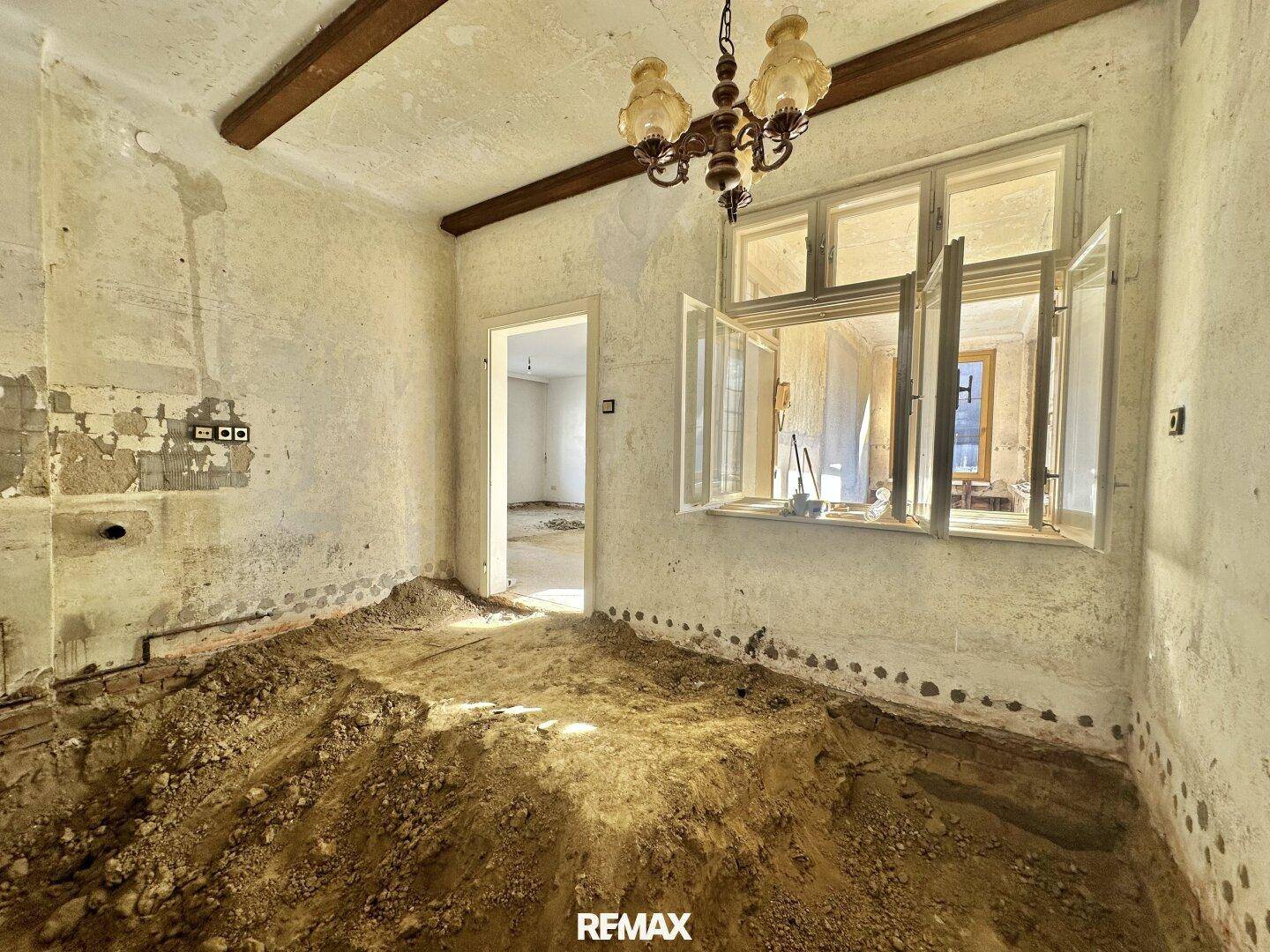 Raum mit sichtbaren Holzbalken, Kronleuchter und großem Fenster, der sich in einem stark renovierungsbedürftigen Zustand befindet.