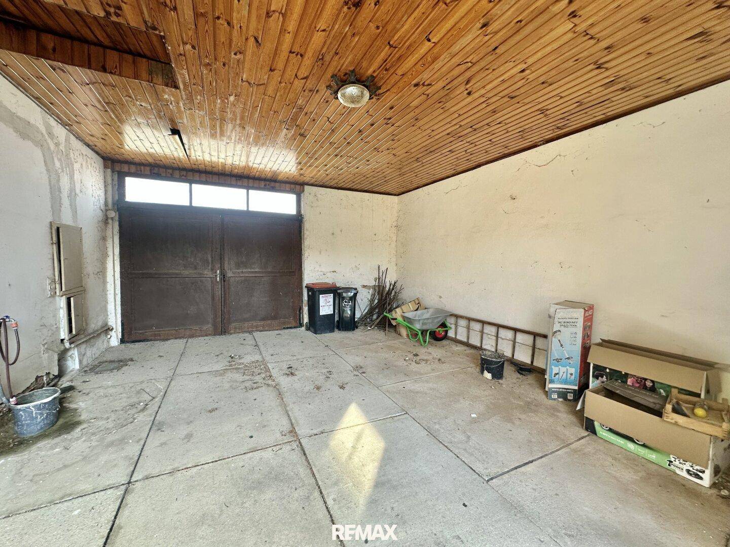 Innenansicht einer Garage mit Holzbalkendecke, Betonboden und großen Doppeltoren, die als Lagerraum dient.