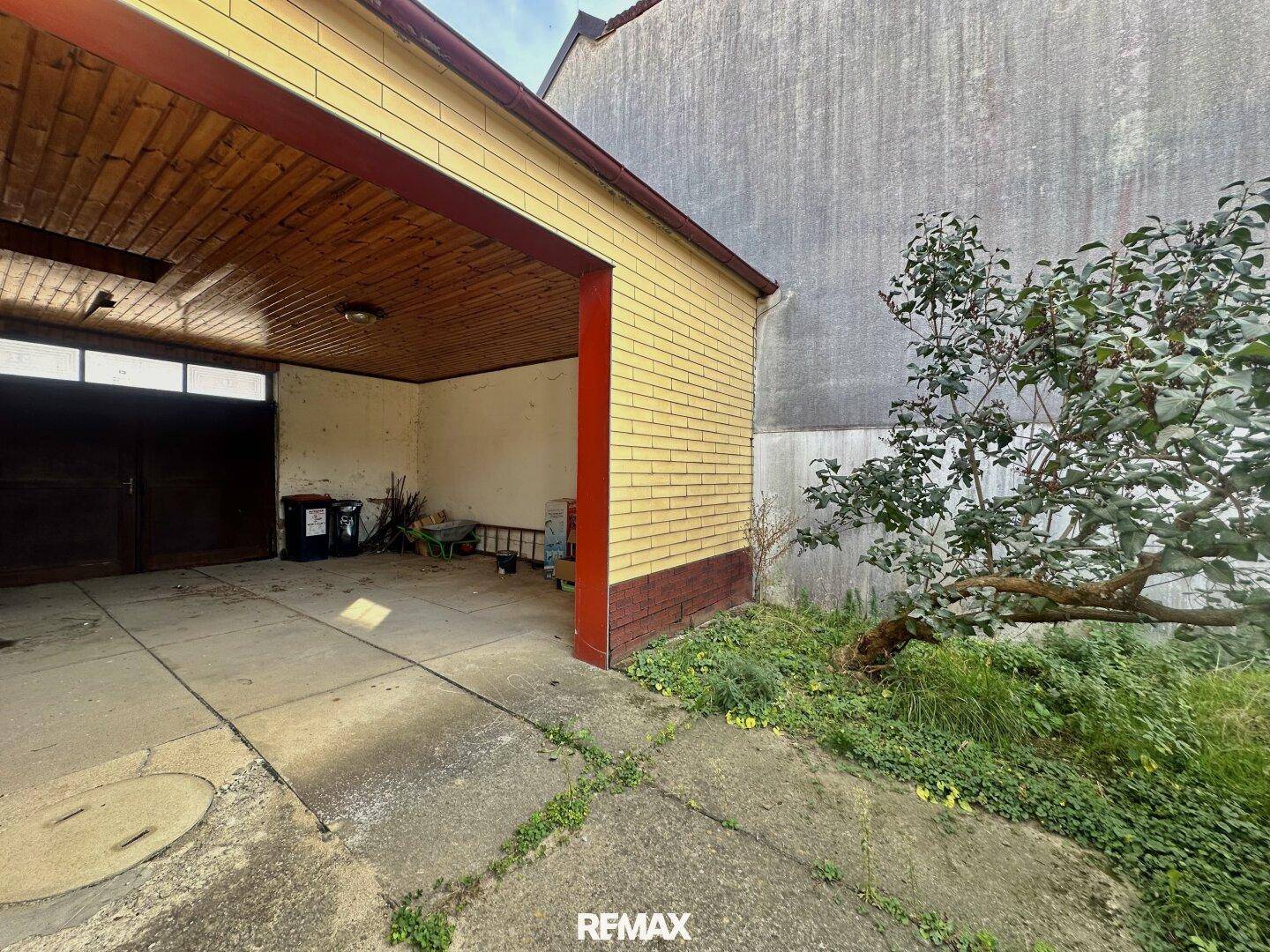 Überdachter Garagenbereich mit Betonboden und Blick auf den verwilderten Garten und eine hohe Mauer.