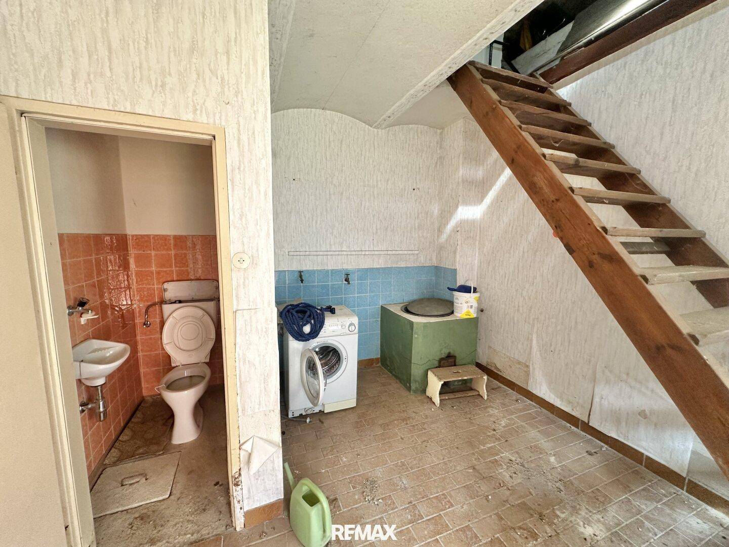 Hauswirtschaftsraum im Keller mit Toilette, Waschbecken, Waschmaschine und Holztreppe, die nach oben führt.