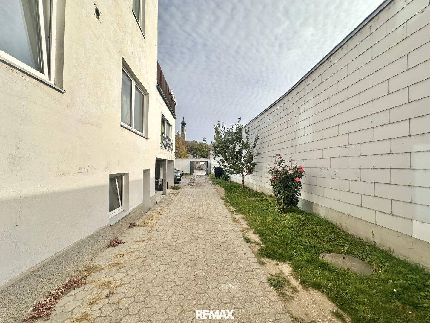 Gepflasterter Weg zwischen Gebäuden mit Grünflächen und einem Baum unter bewölktem Himmel.