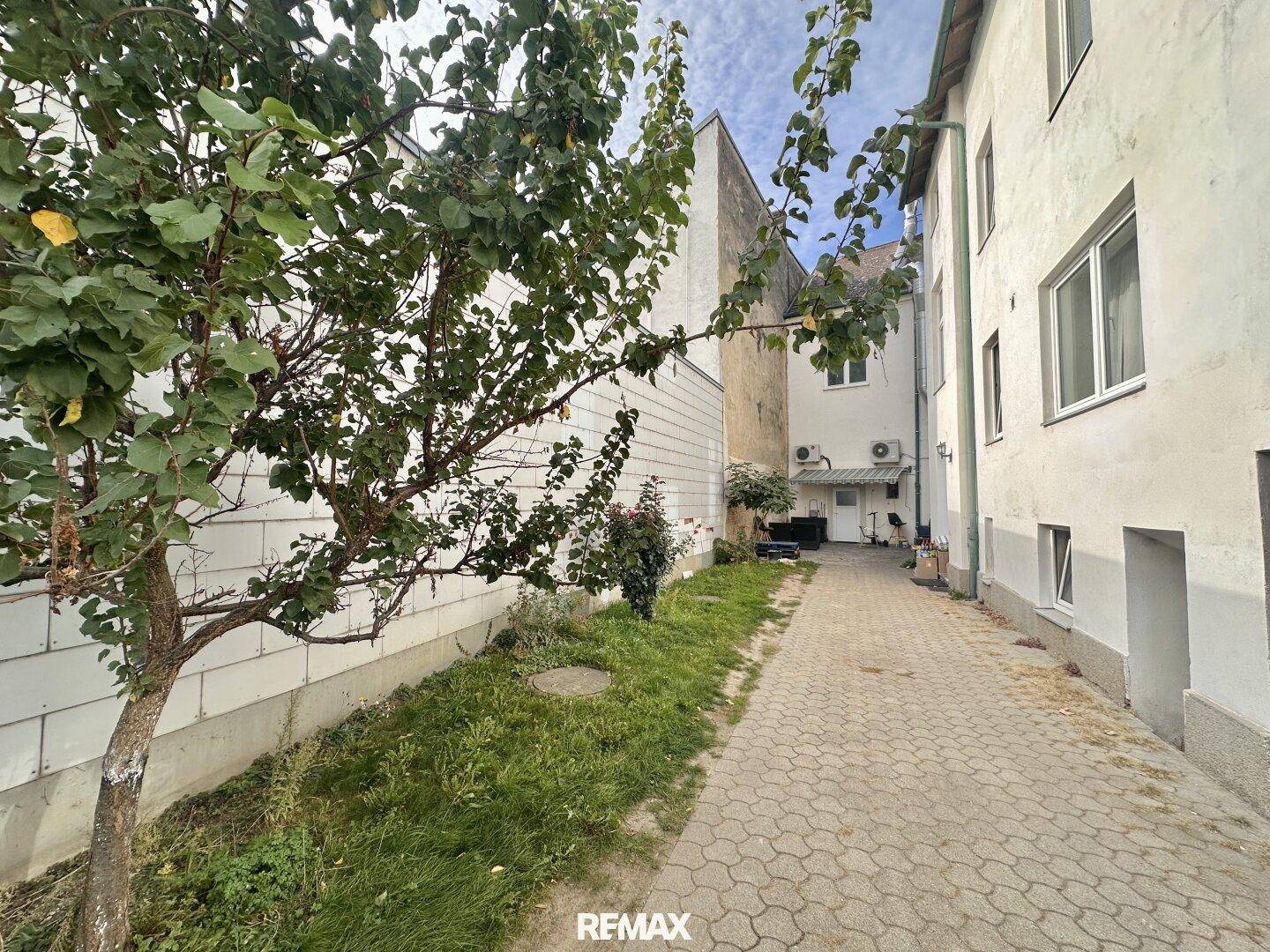 Hinterhofbereich mit gepflastertem Weg, Bäumen und Gebäuden unter bewölktem Himmel.