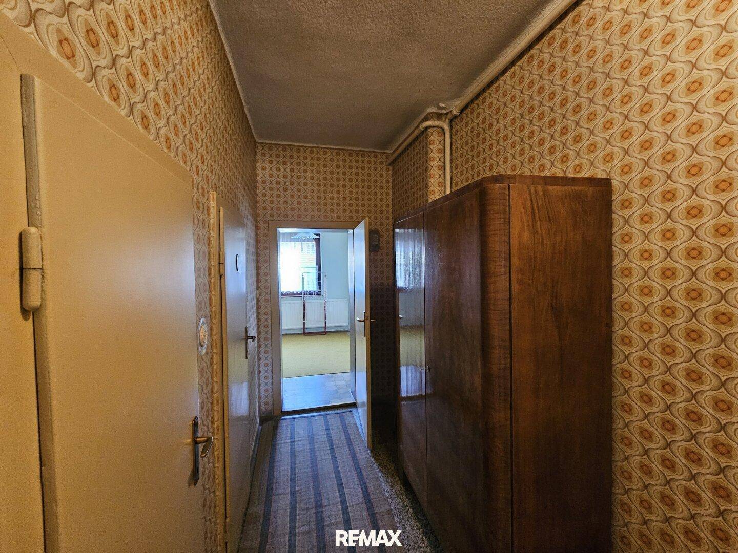 Langer Flur mit gemusterten Tapeten, Holzschrank und Blick in einen weiteren Raum.