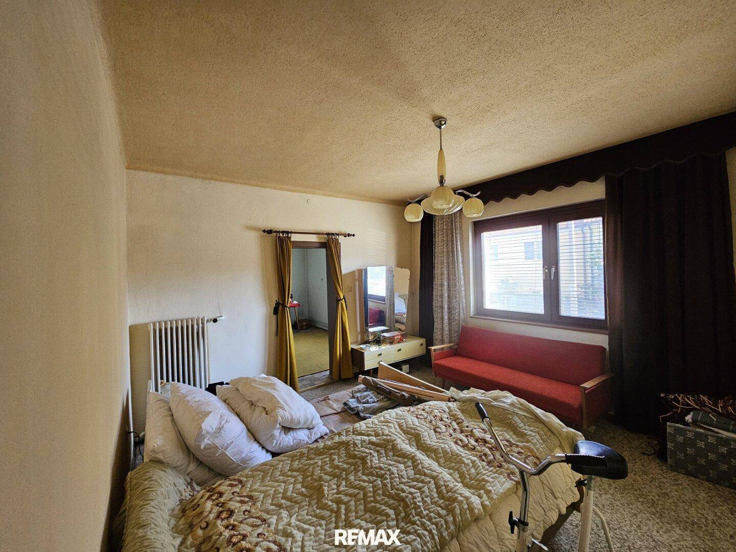 Schlafzimmer mit Bett, rotem Sofa, Fenster und Teppichboden im Originalzustand.