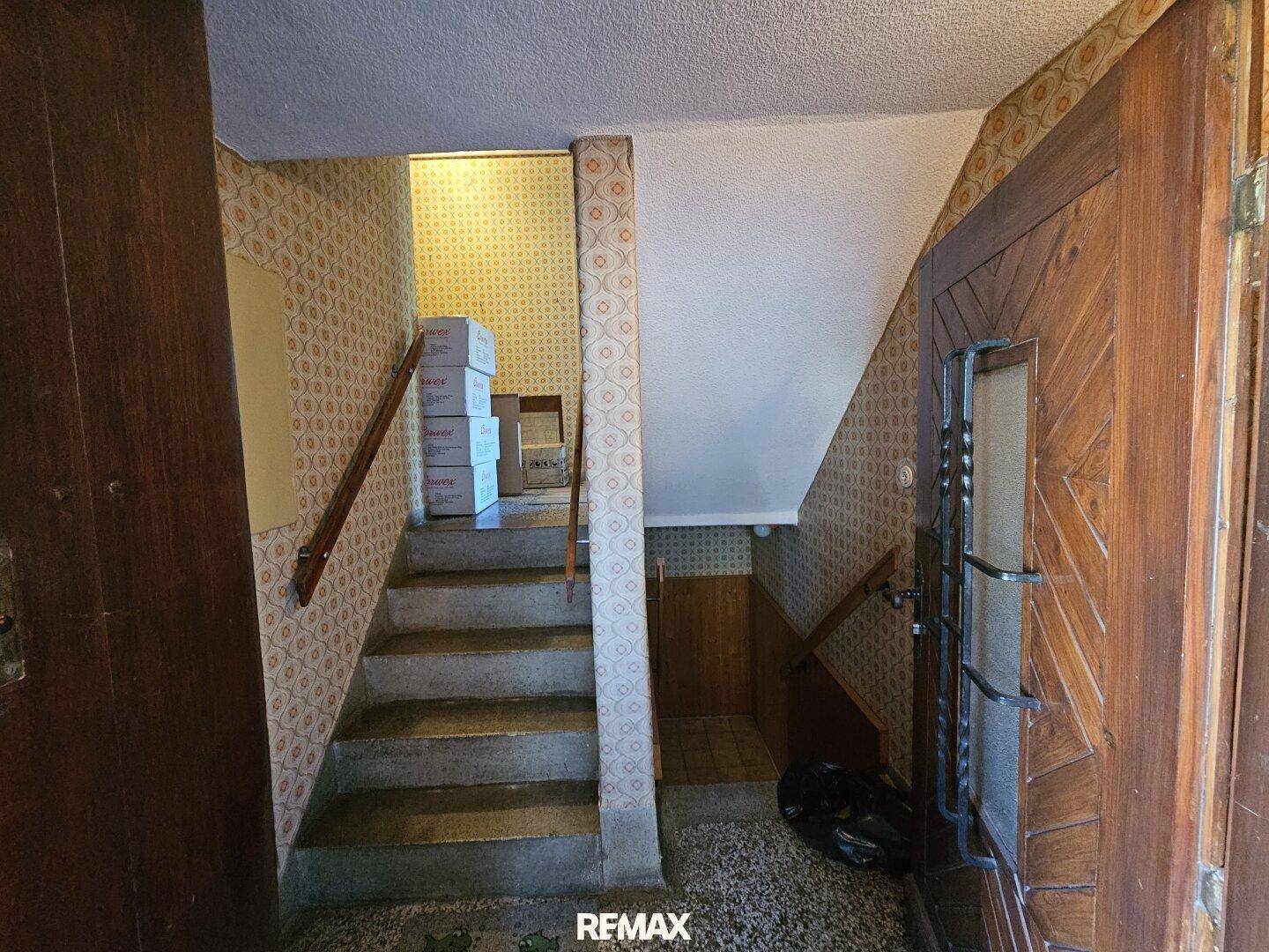Eingangsbereich mit Treppe, gemusterten Tapeten und Holztür im Originalzustand.