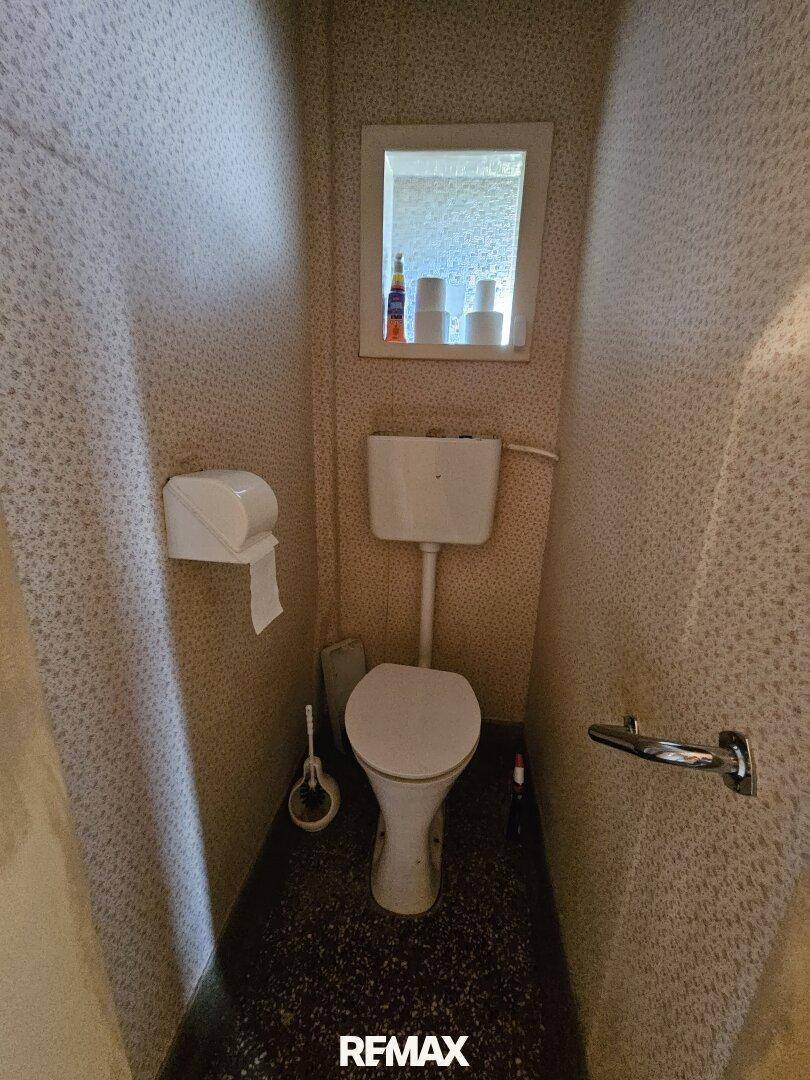 Kleines Gäste-WC mit Toilette, Fenster und gemusterten Tapeten im Originalzustand.