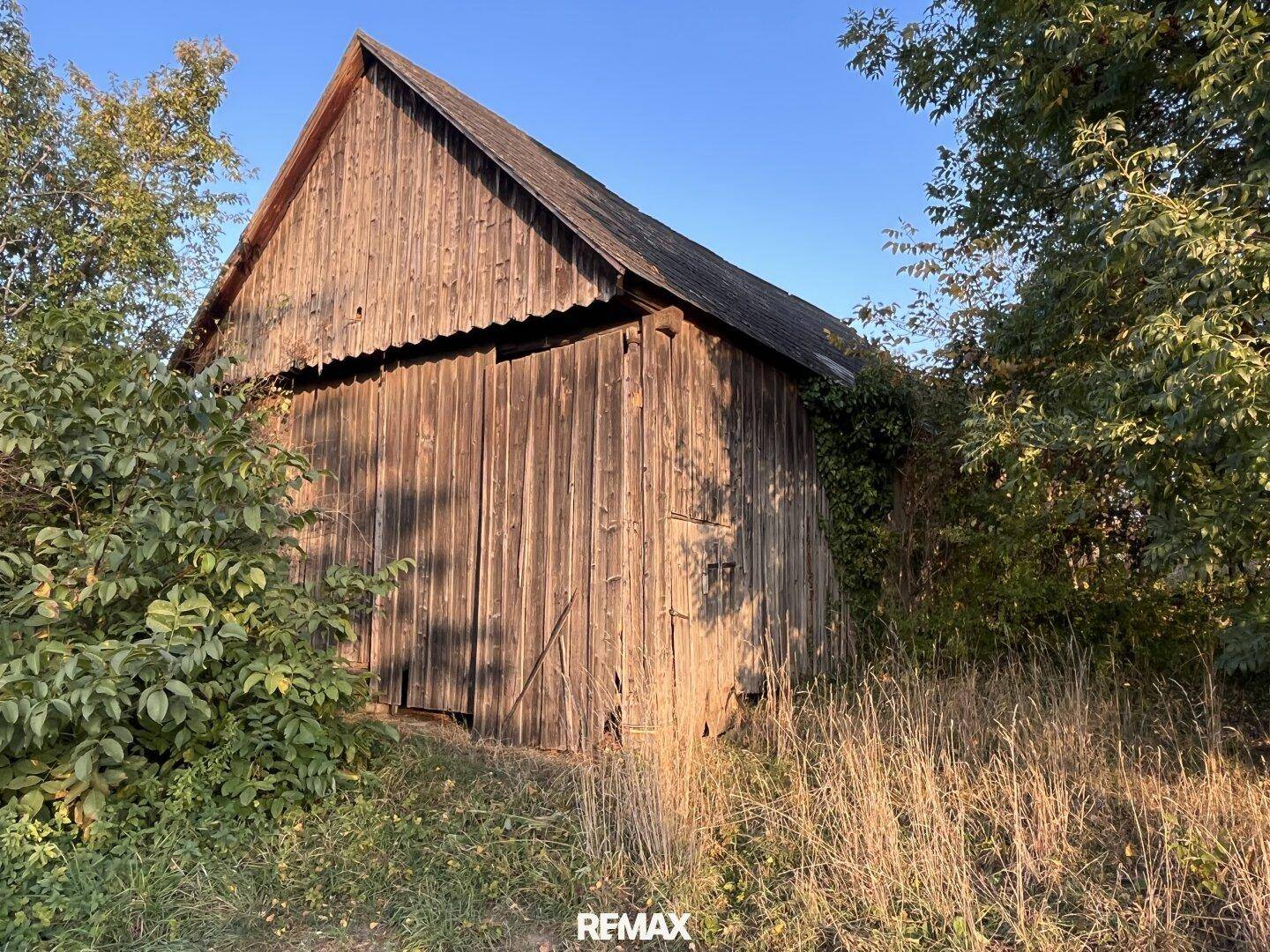 Rustikale Holzscheune mit verwitterten Brettern und einem Giebeldach, umgeben von Vegetation.