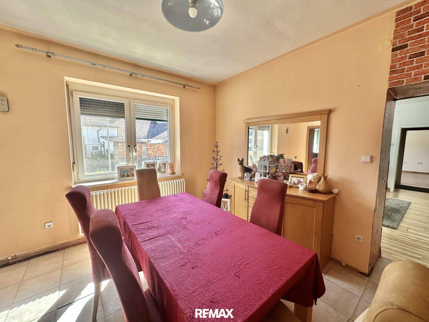 Gemütlicher Essbereich mit Holztisch, roter Tischdecke und Blick nach draußen durch das Fenster.