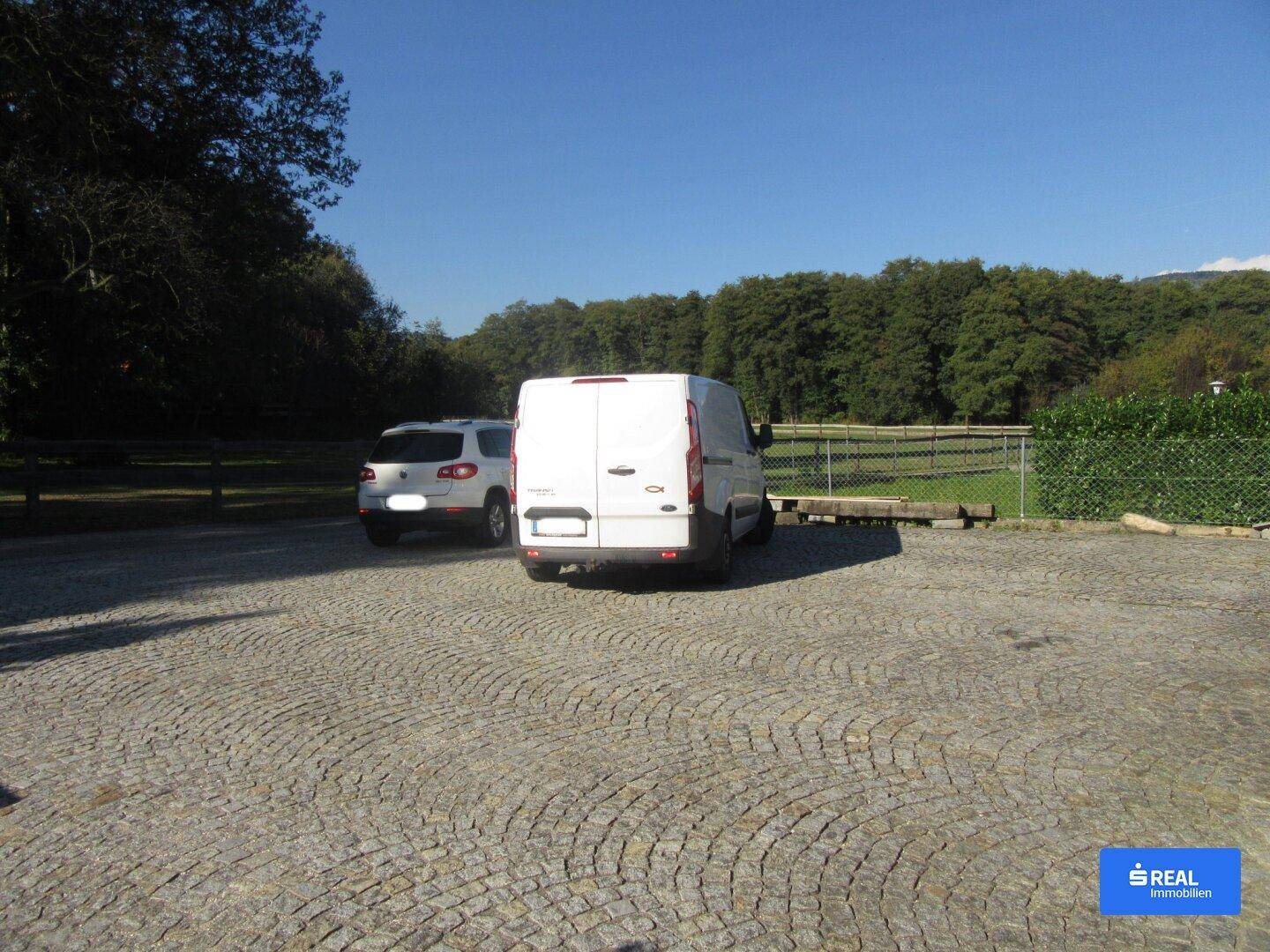 Außenparkplatz mit Kopfsteinpflaster und zwei Fahrzeugen, umgeben von Bäumen und Grünflächen.