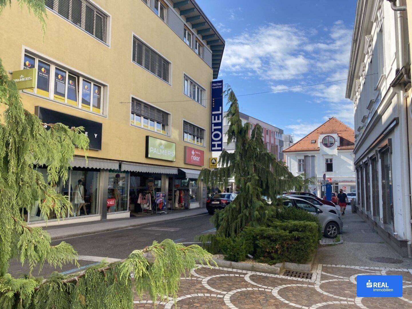 Belebtes Stadtbild mit Geschäften, parkenden Autos und einem Hotel unter blauem Himmel.
