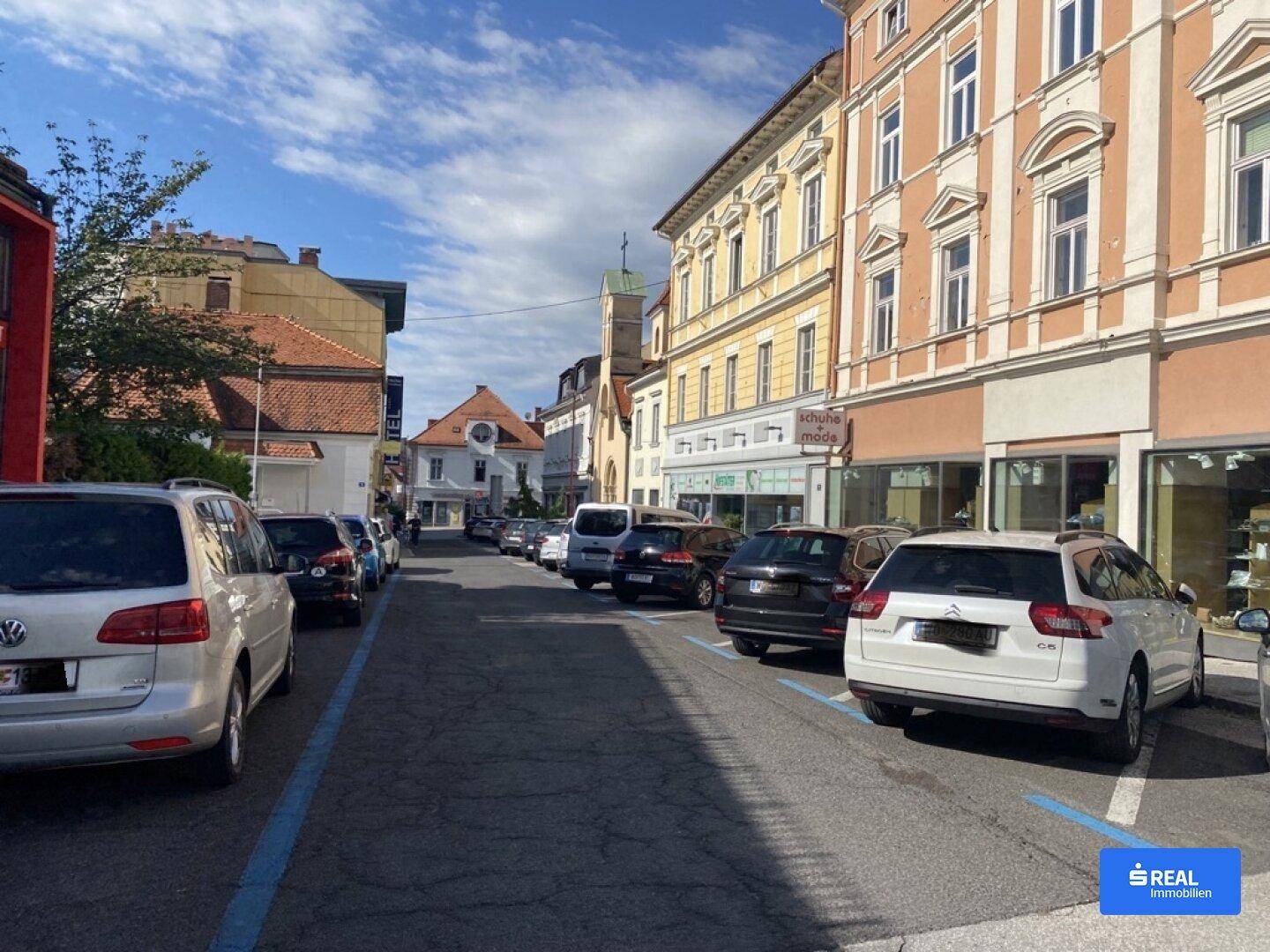 Historische Straße mit geparkten Autos, traditionellen Gebäuden und Geschäften unter blauem Himmel.