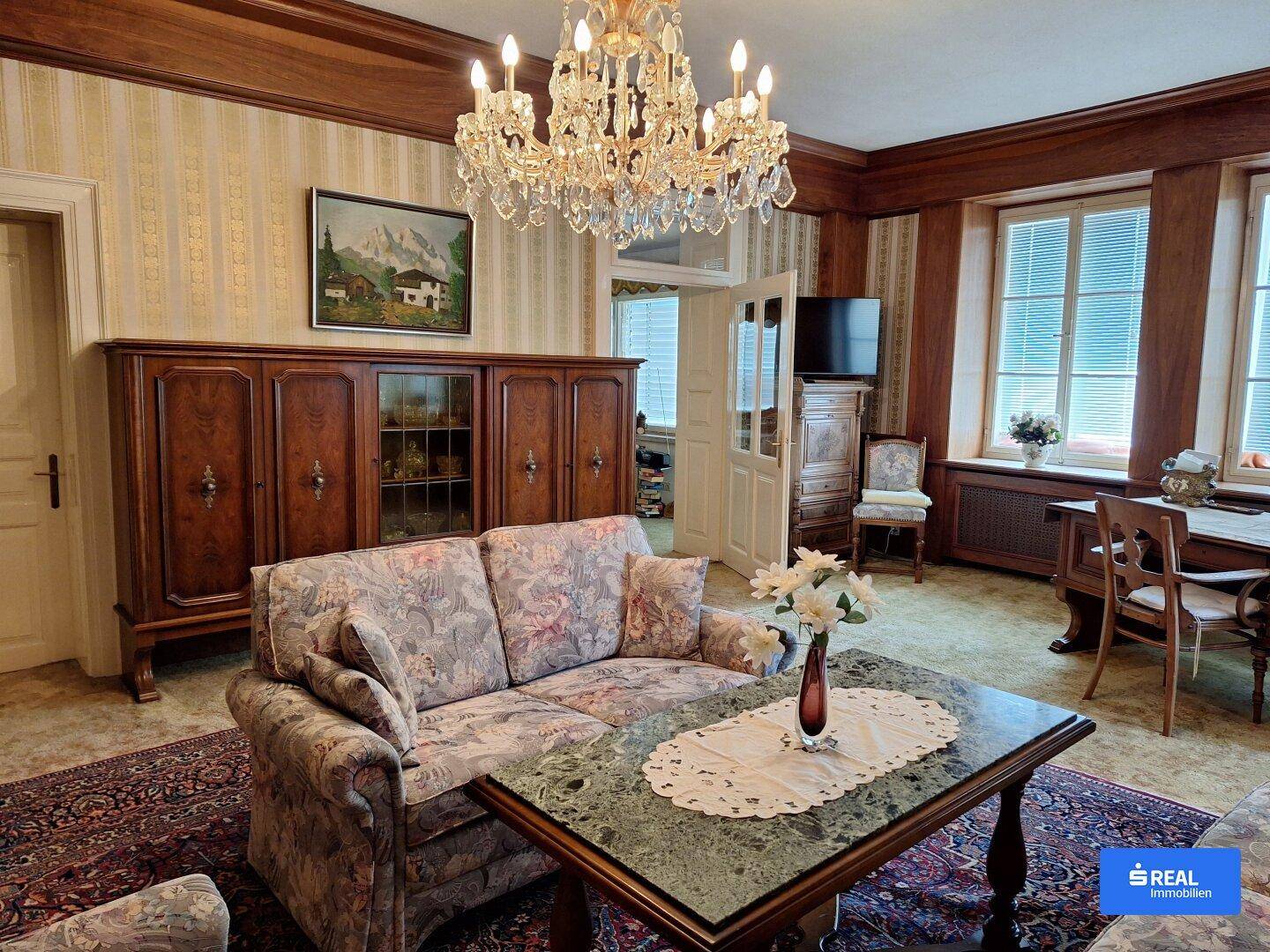 Traditionelles Wohnzimmer mit opulentem Kronleuchter, gemusterten Tapeten und antiken Holzmöbeln.