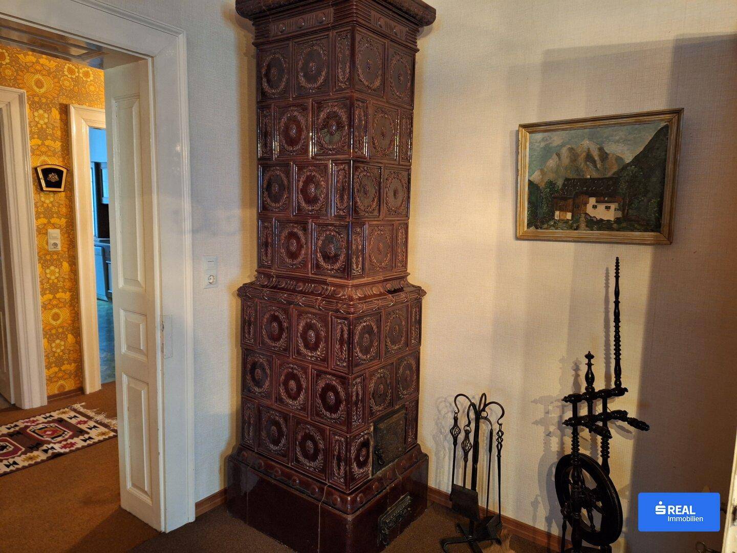 Hoher, verzierter Kachelofen in einem Flur mit gemusterter Tapete und traditioneller Einrichtung.