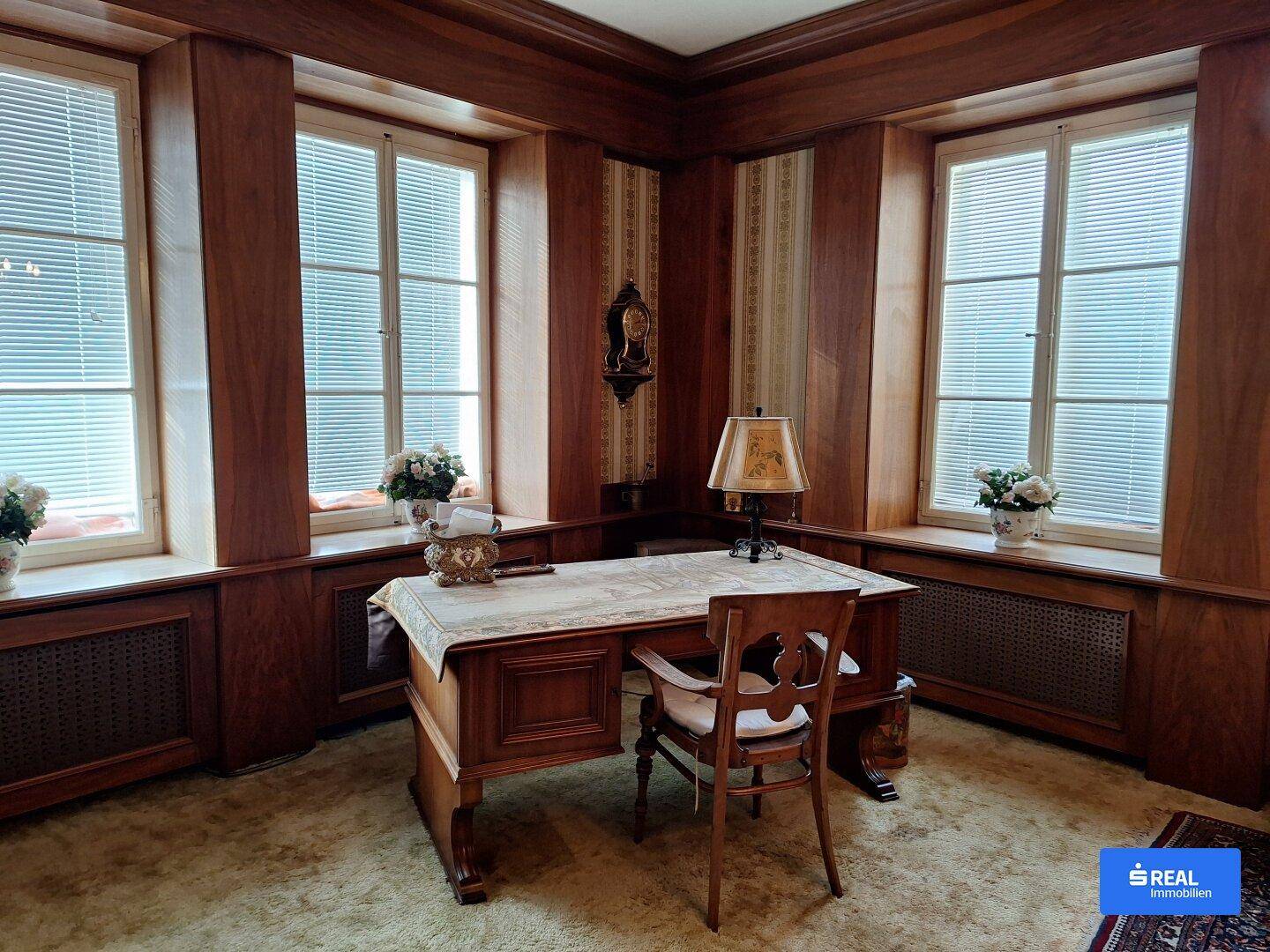 Klassisches Arbeitszimmer mit dunkler Holzvertäfelung, Schreibtisch und großen Fenstern mit Jalousien.