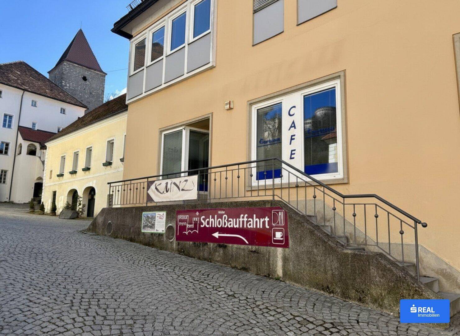 Außenansicht eines Gebäudes mit Café-Eingang, Treppe und Hinweisschildern zur Schlossauffahrt.