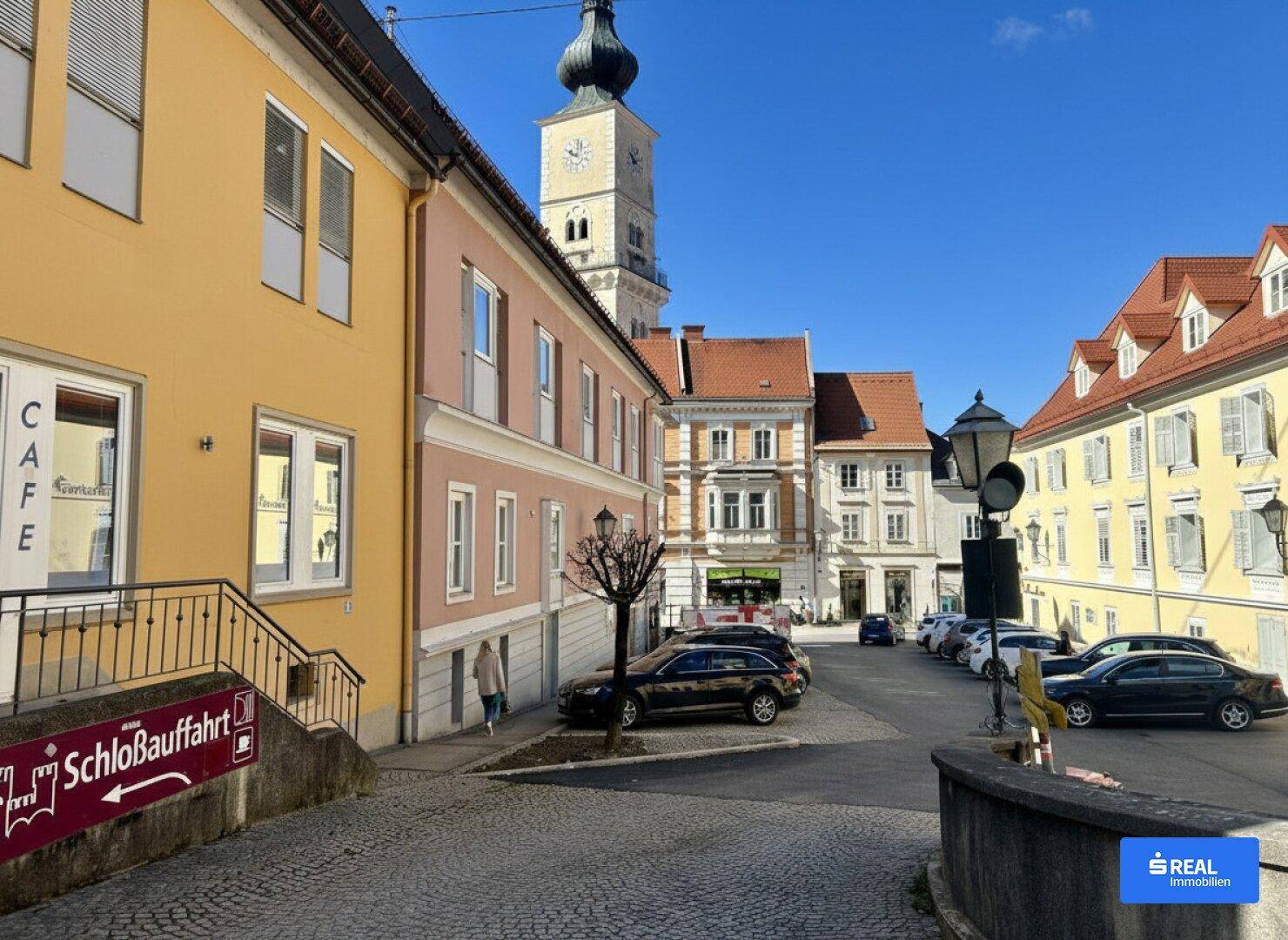 Weitläufige Straßenansicht mit historischen Gebäuden, geparkten Autos und einem markanten Kirchturm im Hintergrund.