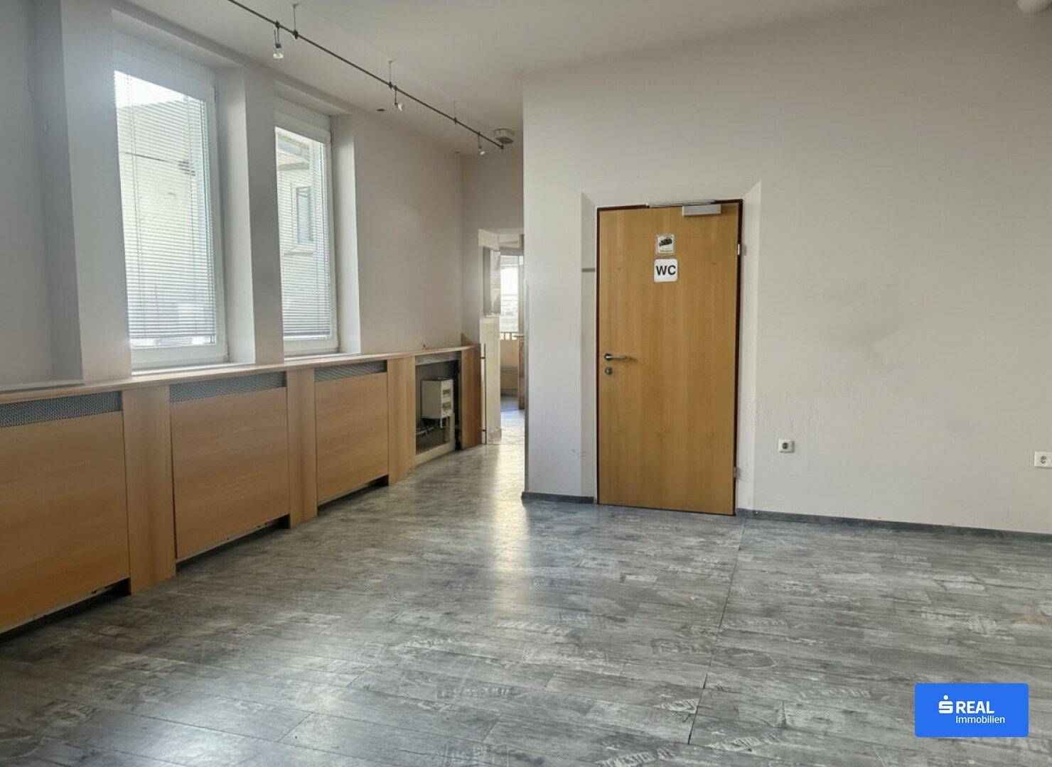 Innenraum mit großen Fenstern, Holzverkleidungen und einer Tür, die zu einer Toilette führt.