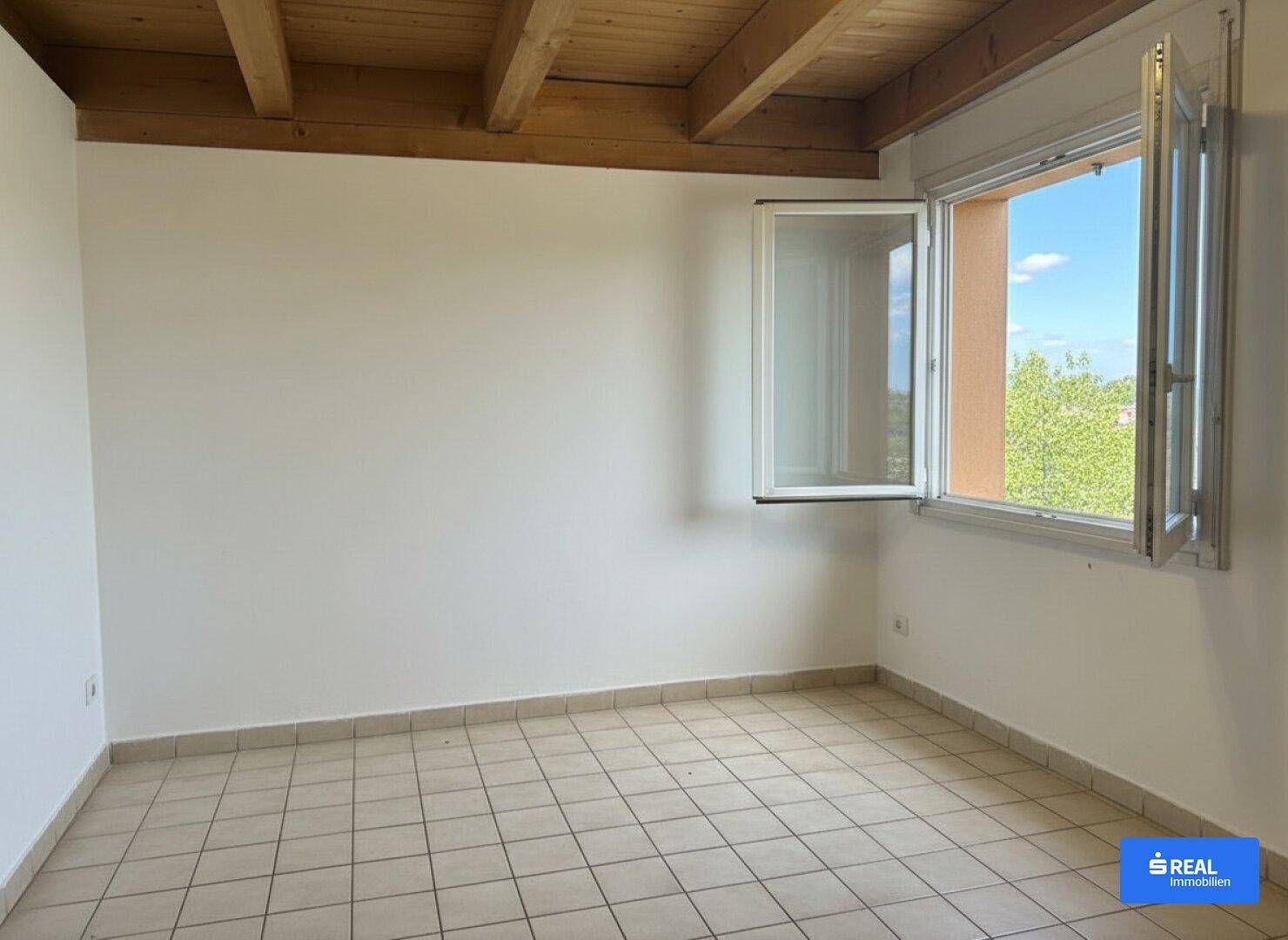 Leerer Raum mit Fliesenboden, weiß gestrichenen Wänden und einem Fenster mit Ausblick ins Grüne.