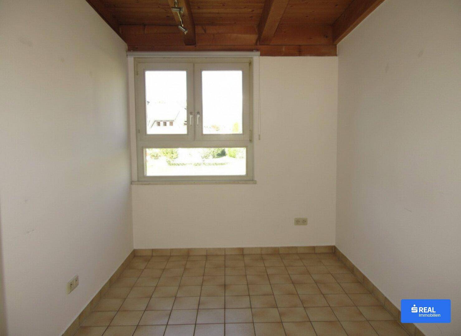 Heller, leerer Raum mit Fliesenboden, Holzbalkendecke und einem großen Fenster mit Ausblick.