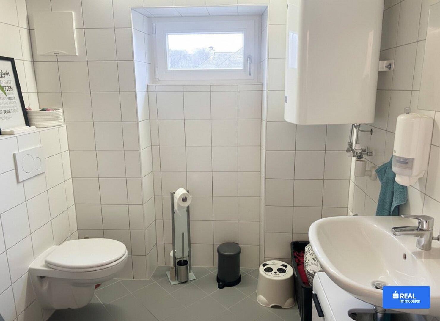 Kleines Badezimmer mit weißen Fliesen, wandhängendem WC, Waschbecken und einem Fenster.