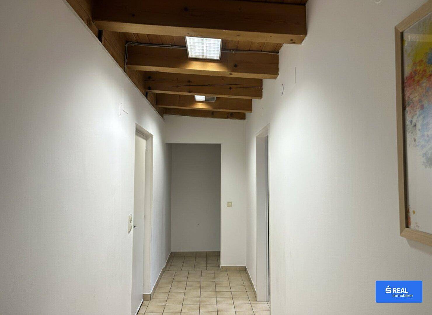 Langer Flur mit Holzbalkendecke, weißen Wänden und Fliesenboden, der zu weiteren Räumen führt.
