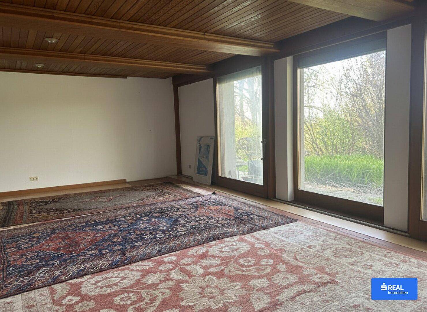 Helles Wohnzimmer mit Holzbalkendecke, großen Fenstern und Zugang zum Außenbereich.