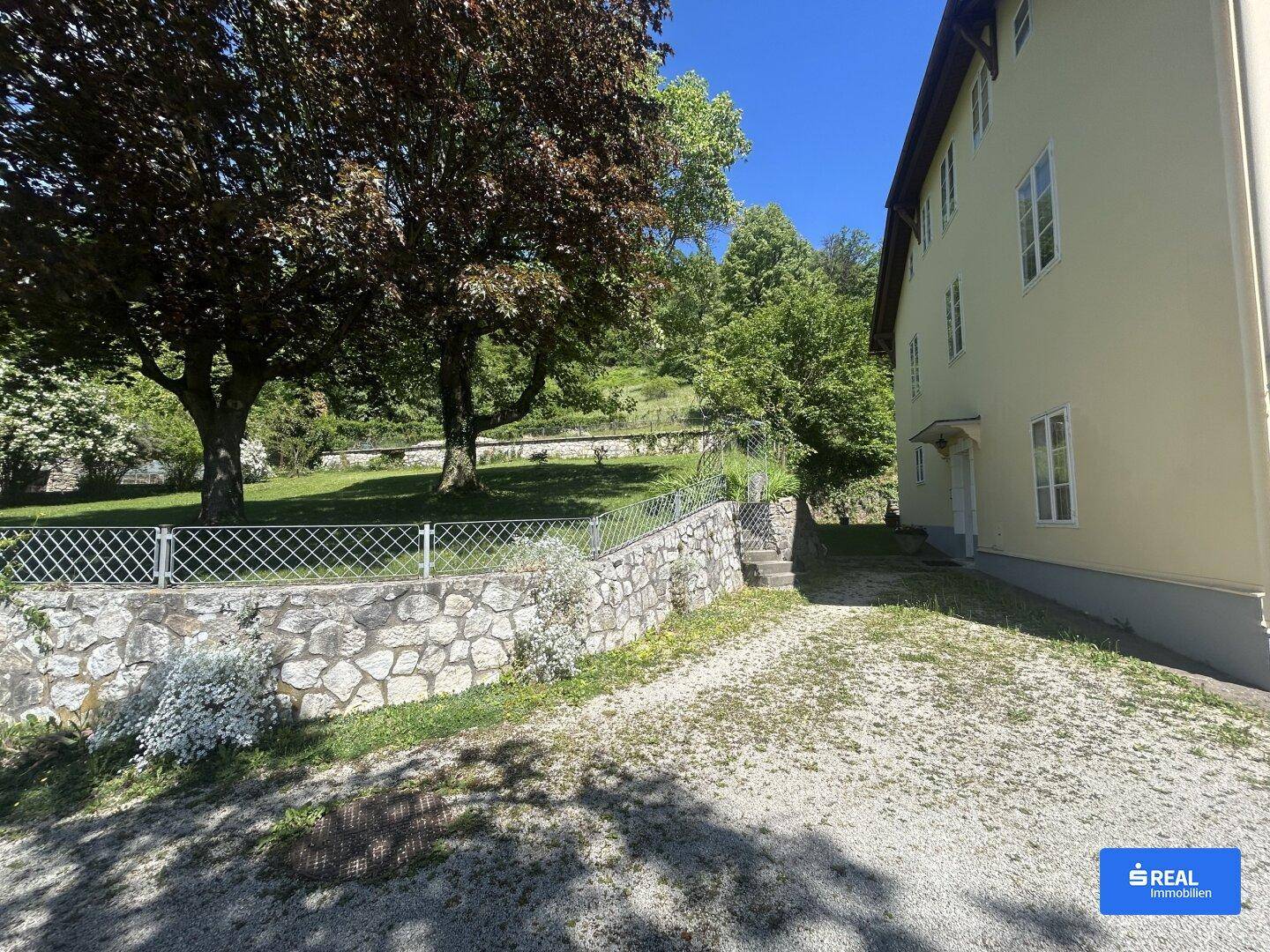 Seitenansicht des Gebäudes mit gelber Fassade, umgeben von einer Steinmauer und üppiger Vegetation.