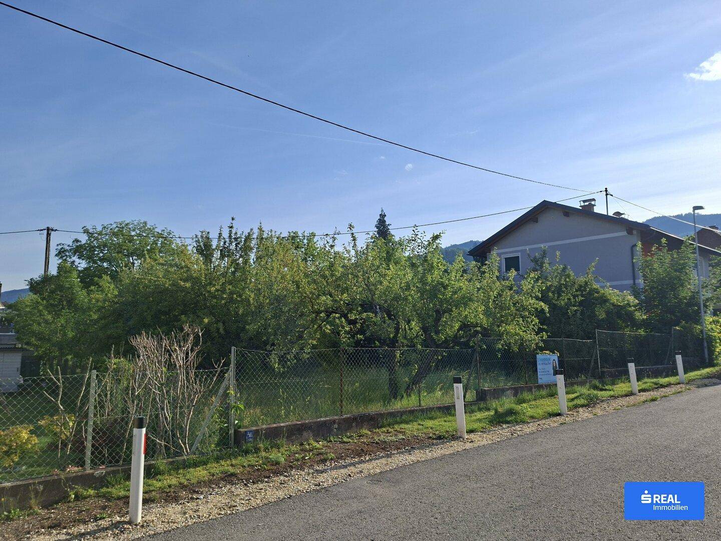 Weitläufige Ansicht des Grundstücks von der Straße mit Wohnhaus, Bäumen und Bergen im Hintergrund.