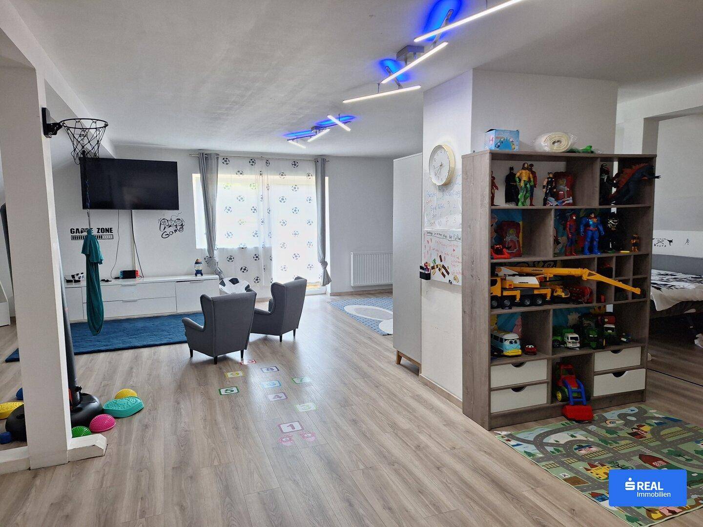 Geräumiger Spielbereich mit Laminatboden, Fernseher und Spielzeugregal.