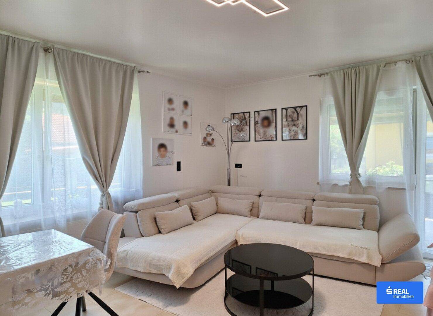 Gemütliches Wohnzimmer mit großem Ecksofa, hellen Vorhängen und Teppichboden.
