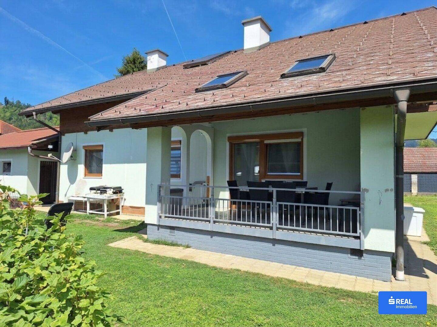 Hausfassade mit überdachter Terrasse, Gartenmöbeln und Blick ins Grüne unter blauem Himmel.